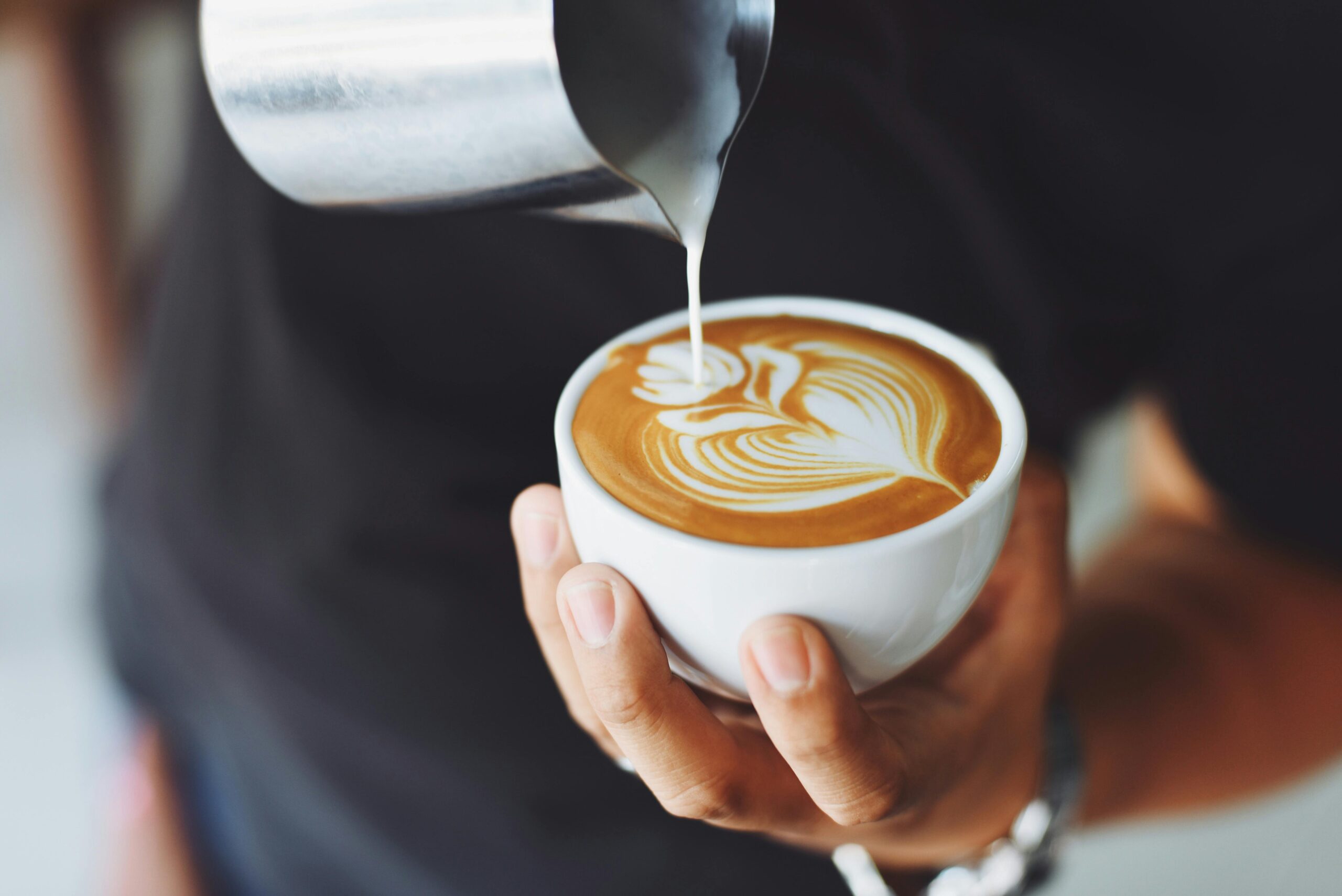 hands of a barista one holding a small white coffee cup and using the other hand to create a flower design with milk